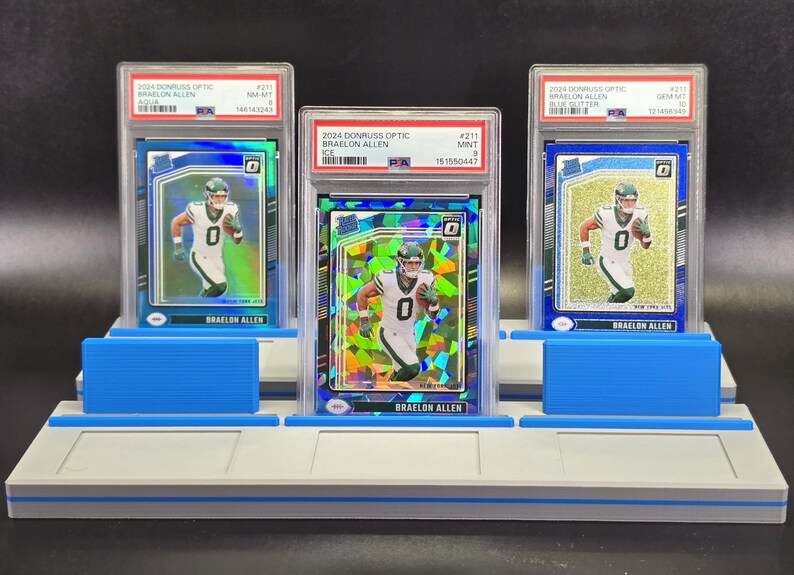 May include: Three graded 2024 Donruss Optic Braelon Allen football cards in PSA plastic cases, displayed on a light grey tiered stand with blue accents. The cards feature the athlete in a white and green uniform, with aqua, ice, and blue glitter finishes.