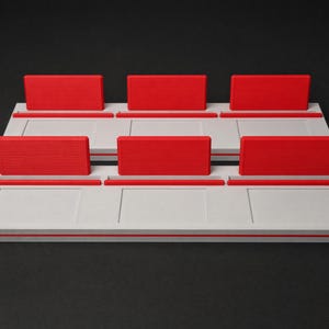 May include: Two rectangular white display stands, each featuring three distinct slots. Each slot is equipped with a vibrant red upright rectangular divider. The stands have a smooth, flat base with recessed panel detailing on the front surface.