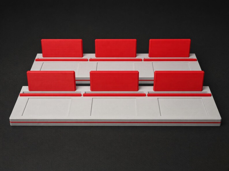 May include: Two rectangular white display stands, each featuring three distinct slots. Each slot is equipped with a vibrant red upright rectangular divider. The stands have a smooth, flat base with recessed panel detailing on the front surface.