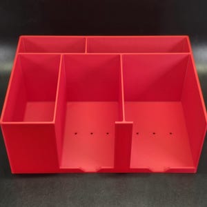 May include: A bright red desk organizer featuring a rectangular shape with five distinct compartments. The unit includes two horizontal slots at the back and three vertical slots at the front, with small drainage holes visible on the base of each section.