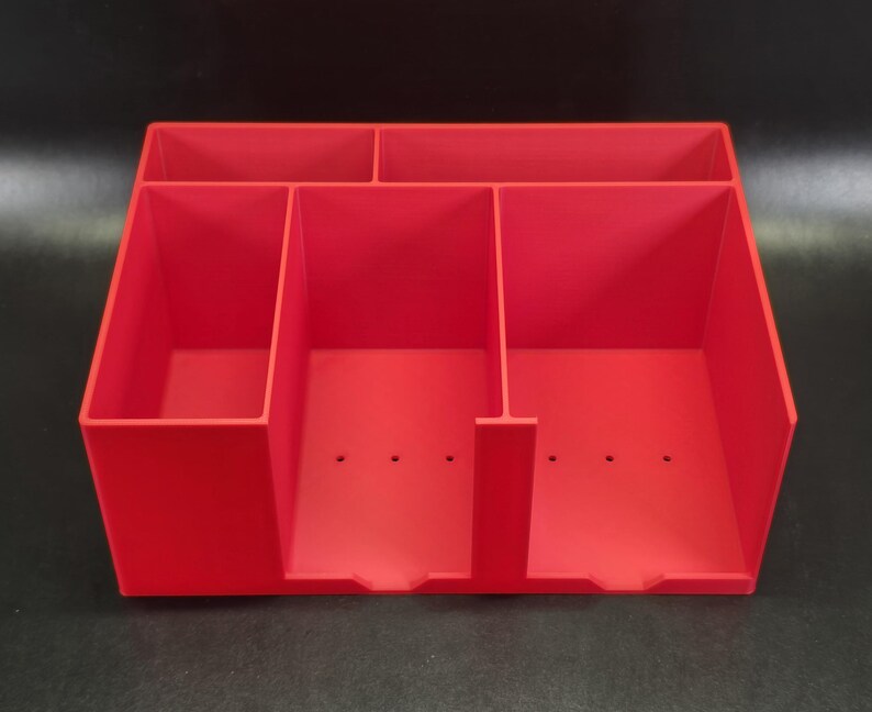 May include: A bright red desk organizer featuring a rectangular shape with five distinct compartments. The unit includes two horizontal slots at the back and three vertical slots at the front, with small drainage holes visible on the base of each section.