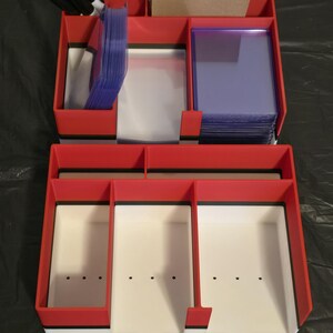 Puede incluir: Un organizador de almacenamiento de m&uacute;ltiples compartimentos en rojo, blanco y negro para tarjetas coleccionables. La secci&oacute;n superior contiene marcadores negros, un trozo de cart&oacute;n y pilas de fundas de pl&aacute;stico transparente.