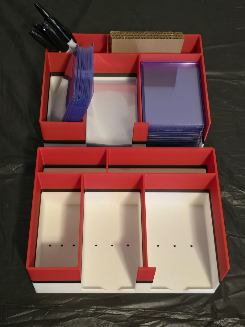 Puede incluir: Un organizador de almacenamiento de m&uacute;ltiples compartimentos en rojo, blanco y negro para tarjetas coleccionables. La secci&oacute;n superior contiene marcadores negros, un trozo de cart&oacute;n y pilas de fundas de pl&aacute;stico transparente.