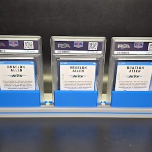 May include: Three Braelon Allen sports trading cards, each encased in a clear PSA protective slab with a white label and QR code. The cards are displayed upright in a grey and blue plastic stand, showing the back of the cards with blue text.