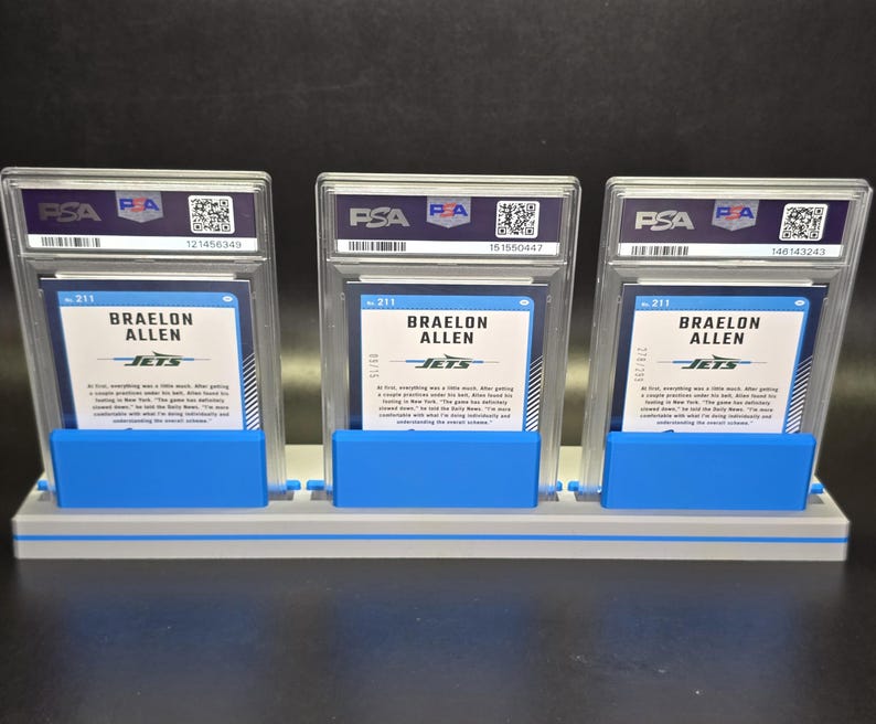 May include: Three Braelon Allen sports trading cards, each encased in a clear PSA protective slab with a white label and QR code. The cards are displayed upright in a grey and blue plastic stand, showing the back of the cards with blue text.