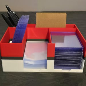 Puede incluir: Un organizador de almacenamiento rojo, blanco y negro con varios compartimentos y ranuras. El organizador contiene pilas de fundas de pl&aacute;stico transparente para tarjetas, una tarjeta rectangular marr&oacute;n y varios bol&iacute;grafos negros.