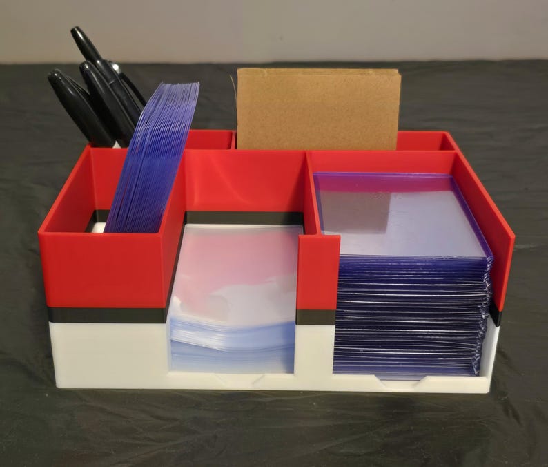 Puede incluir: Un organizador de almacenamiento rojo, blanco y negro con varios compartimentos y ranuras. El organizador contiene pilas de fundas de pl&aacute;stico transparente para tarjetas, una tarjeta rectangular marr&oacute;n y varios bol&iacute;grafos negros.