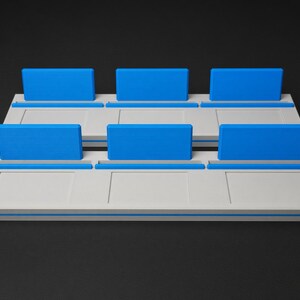 May include: A white rectangular display stand featuring two tiers, each with three distinct slots. Each slot is equipped with a bright blue upright back panel and a matching blue base accent, creating a structured organiser for small items.