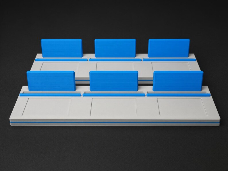 May include: A white rectangular display stand featuring two tiers, each with three distinct slots. Each slot is equipped with a bright blue upright back panel and a matching blue base accent, creating a structured organiser for small items.