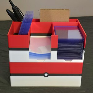 Puede incluir: Un organizador de escritorio de pl&aacute;stico rojo, blanco y negro con m&uacute;ltiples compartimentos. Contiene marcadores negros, una pila de fundas para tarjetas de pl&aacute;stico transparente azulado, un inserto de cart&oacute;n marr&oacute;n y tarjeteros r&iacute;gidos.