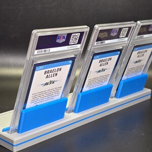 May include: Three Braelon Allen sports trading cards in clear PSA protective slabs, displayed upright in a light grey and blue plastic stand. Each card features the player's name, the Jets team logo, and text on the back, with a PSA logo at the top.