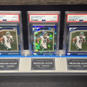 May include: Three 2024 Donruss Optic Braelon Allen trading cards in PSA graded slabs, displayed on a light grey stand. The cards feature the athlete in a white New York Jets uniform, labelled as Aqua, Ice, and Blue Glitter parallels with PSA grades 8, 9, and 10.