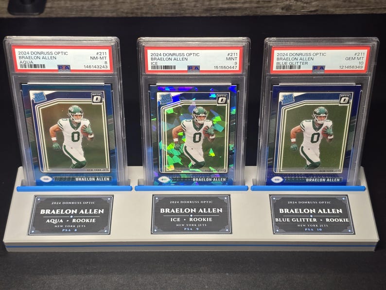 May include: Three 2024 Donruss Optic Braelon Allen trading cards in PSA graded slabs, displayed on a light grey stand. The cards feature the athlete in a white New York Jets uniform, labelled as Aqua, Ice, and Blue Glitter parallels with PSA grades 8, 9, and 10.