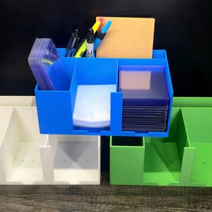 May include: Three rectangular plastic desk organizers in white, blue, and green. Each unit features multiple vertical compartments for storage. The blue organizer is filled with clear plastic card sleeves, pens, and a tan notepad.