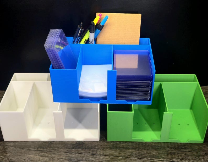 May include: Three rectangular plastic desk organizers in white, blue, and green. Each unit features multiple vertical compartments for storage. The blue organizer is filled with clear plastic card sleeves, pens, and a tan notepad.