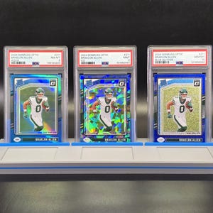 May include: Three graded 2024 Donruss Optic Braelon Allen football cards in PSA plastic cases, displayed on a grey stand. The cards feature the player in a white and green uniform, with distinct aqua, ice, and blue glitter reflective card finishes.