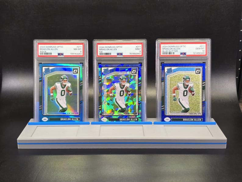 May include: Three graded 2024 Donruss Optic Braelon Allen football cards in PSA plastic cases, displayed on a grey stand. The cards feature the player in a white and green uniform, with distinct aqua, ice, and blue glitter reflective card finishes.