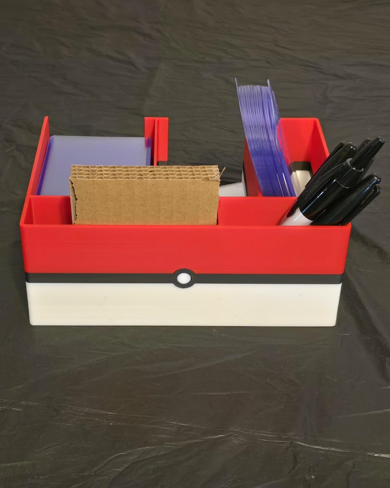 Puede incluir: Un organizador de escritorio de pl&aacute;stico rojo, blanco y negro con una franja horizontal y un emblema circular central. El recipiente cuenta con varios compartimentos que contienen fundas de pl&aacute;stico transparentes, cart&oacute;n y marcadores negros.