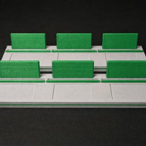May include: A two-tiered display stand featuring a light grey base with rectangular recessed panels and a thin green stripe along the front edge. Each tier includes three upright green rectangular dividers and three horizontal green slots.