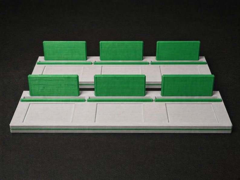 May include: A two-tiered display stand featuring a light grey base with rectangular recessed panels and a thin green stripe along the front edge. Each tier includes three upright green rectangular dividers and three horizontal green slots.