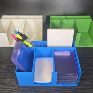 May include: A blue plastic desk organizer containing stacks of clear card sleeves and a white card deck. Behind it sit an off-white organizer and a green organizer. Three markers in pink, yellow, and black are tucked into the blue organizer's side.