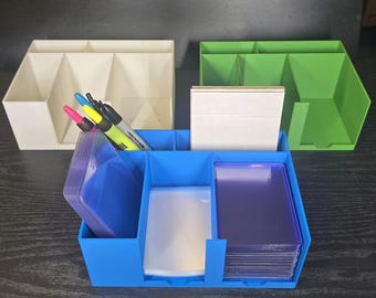 May include: A blue plastic desk organiser containing stacks of clear card sleeves and a white card deck. Behind it sit an off-white organiser and a green organiser. Three markers in pink, yellow, and black are tucked into the blue organiser's side.