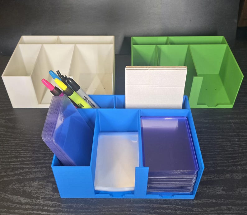 May include: A blue plastic desk organizer containing stacks of clear card sleeves and a white card deck. Behind it sit an off-white organizer and a green organizer. Three markers in pink, yellow, and black are tucked into the blue organizer's side.