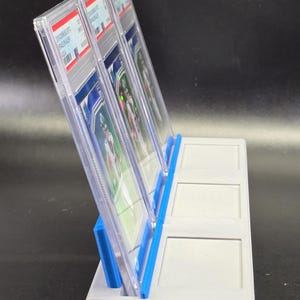 May include: A light grey plastic display stand with blue accents, designed to hold three rectangular trading card slabs in an upright position. The stand features three distinct slots, with two clear plastic slabs currently inserted in the first two slots.