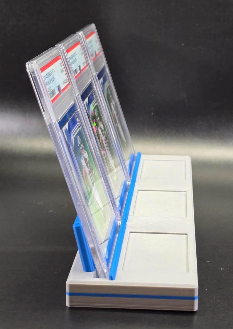 May include: A light grey plastic display stand with blue accents, designed to hold three rectangular trading card slabs in an upright position. The stand features three distinct slots, with two clear plastic slabs currently inserted in the first two slots.