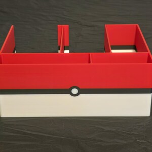 Puede incluir: Un organizador de escritorio rojo, blanco y negro con un dise&ntilde;o horizontal y m&uacute;ltiples compartimentos. El exterior muestra un patr&oacute;n de bloques de color con un emblema circular central, imitando la apariencia de un dispositivo esf&eacute;rico de captura.