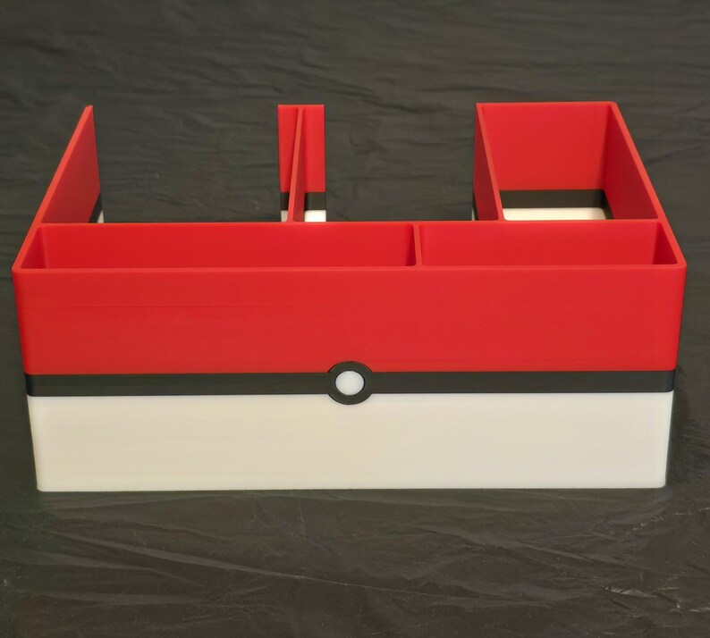 Puede incluir: Un organizador de escritorio rojo, blanco y negro con un dise&ntilde;o horizontal y m&uacute;ltiples compartimentos. El exterior muestra un patr&oacute;n de bloques de color con un emblema circular central, imitando la apariencia de un dispositivo esf&eacute;rico de captura.