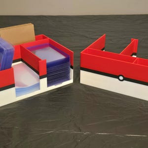 Puede incluir: Dos organizadores de pl&aacute;stico rojo, blanco y negro con una franja negra horizontal y un detalle circular central. El organizador izquierdo contiene pilas de fundas transparentes para tarjetas, un bloque rectangular beige y dos bol&iacute;grafos negros.