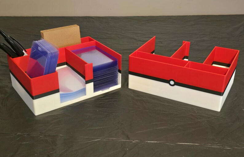Puede incluir: Dos organizadores de pl&aacute;stico rojo, blanco y negro con una franja negra horizontal y un detalle circular central. El organizador izquierdo contiene pilas de fundas transparentes para tarjetas, un bloque rectangular beige y dos bol&iacute;grafos negros.