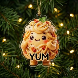 May include: A clear acrylic hanging ornament shaped like a bowl of spaghetti carbonara with a cute cartoon face, pink cheeks, and small arms. The pasta is topped with bacon bits and a sprig of parsley, with the word YUM printed in bold black letters.
