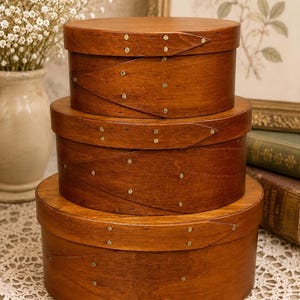 May include: A set of three stacked oval wooden boxes with a medium brown finish. Each box features a lid with a distinctive swallowtail joint secured by small brass-colored metal tacks along the overlapping edges of the bent wood sides.
