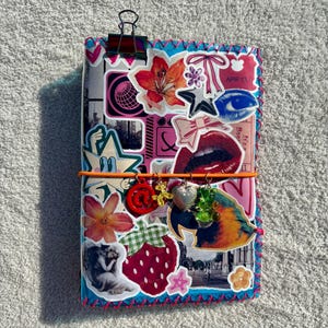May include: A rectangular journal covered in a collage of stickers including flowers, a red lip, a strawberry, and a parrot. An orange elastic band holds four charms: a red at-symbol, a yellow balloon dog, a silver heart, and a green gummy bear.