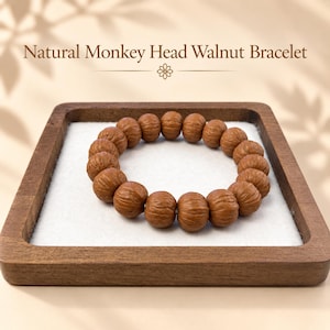 May include: A bracelet composed of round, brown, textured monkey head walnut beads arranged in a circular pattern. The text Natural Monkey Head Walnut Bracelet is displayed above the item, which is resting on a square wooden tray.