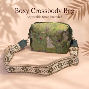 May include: A boxy crossbody bag featuring a printed scene of rabbits in a green meadow with colorful flowers. The bag includes an adjustable strap with a white and green geometric pattern and gold-tone hardware. Text reads: Boxy Crossbody Bag, Adjustable Strap Included.