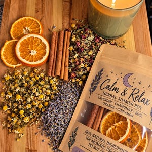 Può includere: Una bustina etichettata Calm & Relax Herbal Simmer Pot contenente fette di arancia essiccate e bastoncini di cannella, insieme a fiori di camomilla, boccioli di lavanda e petali di rosa essiccati. Una candela accesa è posta accanto.