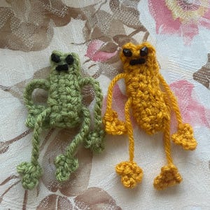 May include: Two small, handmade crochet frog figures, one in sage green and one in mustard yellow. Each frog features a rectangular body, long thin limbs with small feet, and black bead eyes with a simple stitched mouth detail on their faces.