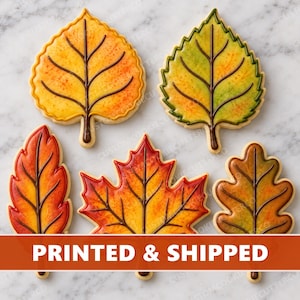May include: Five leaf-shaped cookies decorated with royal icing in autumn colors. The cookies feature detailed brown piped veins and gradients of yellow, orange, green, and red. A prominent orange banner across the bottom displays the text PRINTED & SHIPPED.