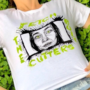 Może przedstawiać: Biała koszulka z zielonym nadrukiem graficznym przedstawiającym twarz kobiety w czarno-białej ramce. Wokół ramki napisano "FETCH THE BODY".