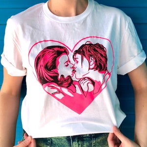May include: White t-shirt with a pink heart graphic featuring a couple kissing. The heart graphic is outlined in black.