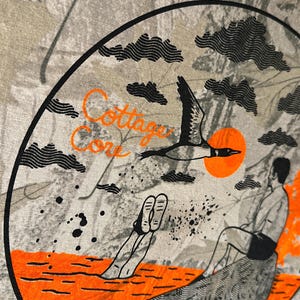 May include: A t-shirt with a circular graphic featuring a bird in flight, an orange sun, and a person sitting on a rock. The words "Cottage Core" are in orange script. The background has a camouflage pattern.