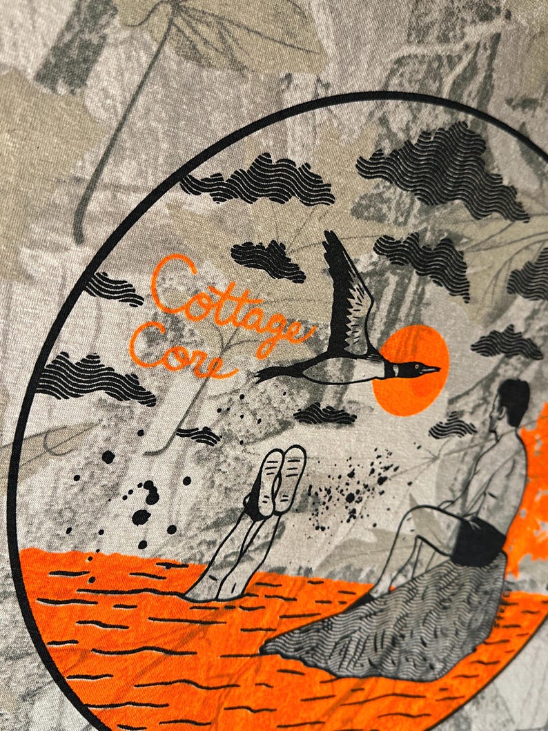 May include: A t-shirt with a circular graphic featuring a bird in flight, an orange sun, and a person sitting on a rock. The words "Cottage Core" are in orange script. The background has a camouflage pattern.