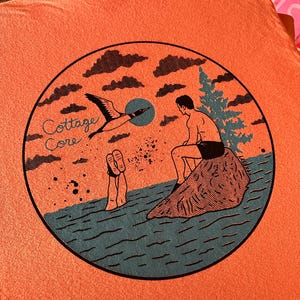 May include: Orange t-shirt with a circular graphic. The graphic features a person sitting on a rock, a bird in flight, and the text "Cottage Core". The design is in shades of blue, orange, and black.