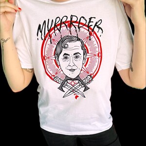 May include: A white t-shirt featuring a graphic print of a person's head centered within a red circular emblem with radiating lines. Below the head are two crossed daggers. The text "MYRRROER" is printed in black above the circular design.