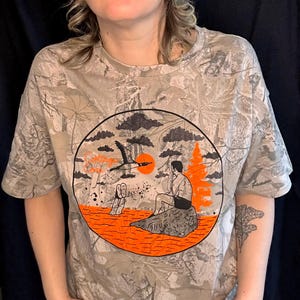May include: A beige camouflage-patterned t-shirt with a circular graphic featuring a bird in flight, a person sitting on a rock, and an orange sun. The shirt has short sleeves.