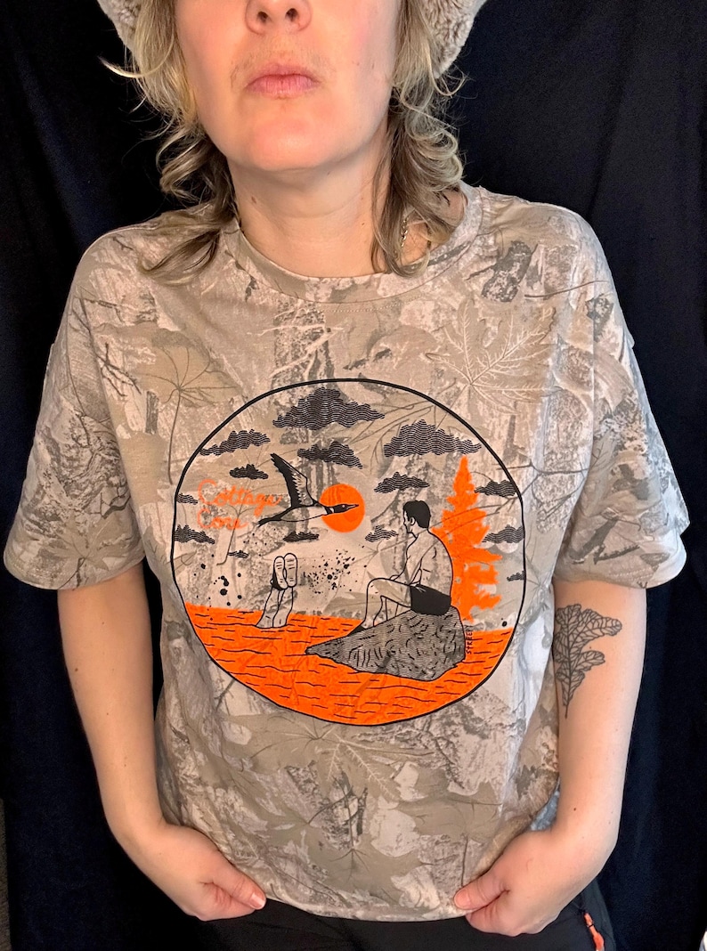 May include: A beige camouflage-patterned t-shirt with a circular graphic featuring a bird in flight, a person sitting on a rock, and an orange sun. The shirt has short sleeves.