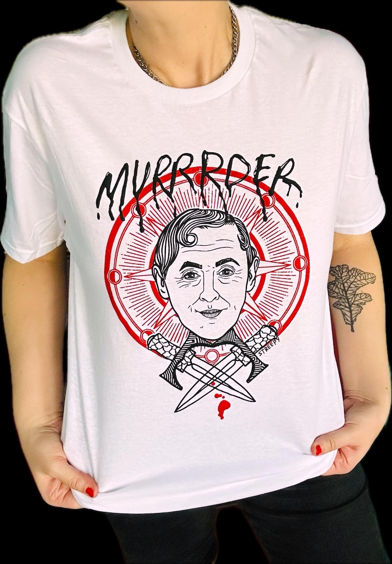 May include: A white t-shirt featuring a graphic print of a person's face centered within a red circular emblem with radiating lines. Below the face are two crossed daggers. The text "MYRRRDER" is printed in a black, dripping font above the emblem.
